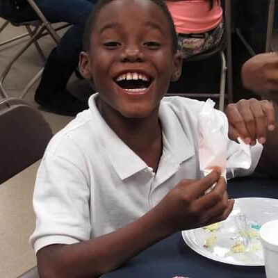 A young boy smiles while eating at a table.